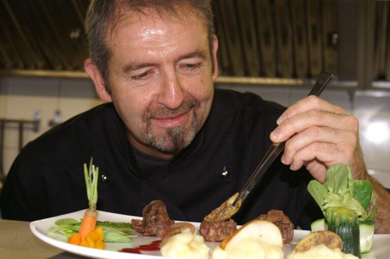 Ein Koch in schwarzer Kleidung richtet konzentriert ein buntes Gericht mit Pinzette auf einem Teller an.