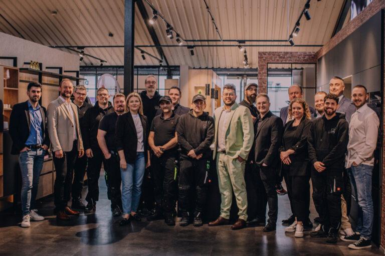 Eine Gruppe von 18 Personen in formeller Kleidung posiert lächelnd für ein Foto in einem modernen Raum.