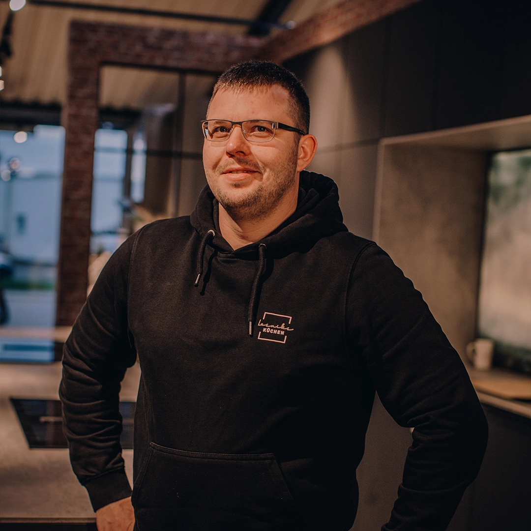 Person im schwarzen Hoodie lächelt in moderner Küche; warme Beleuchtung schafft gemütliche Atmosphäre.
