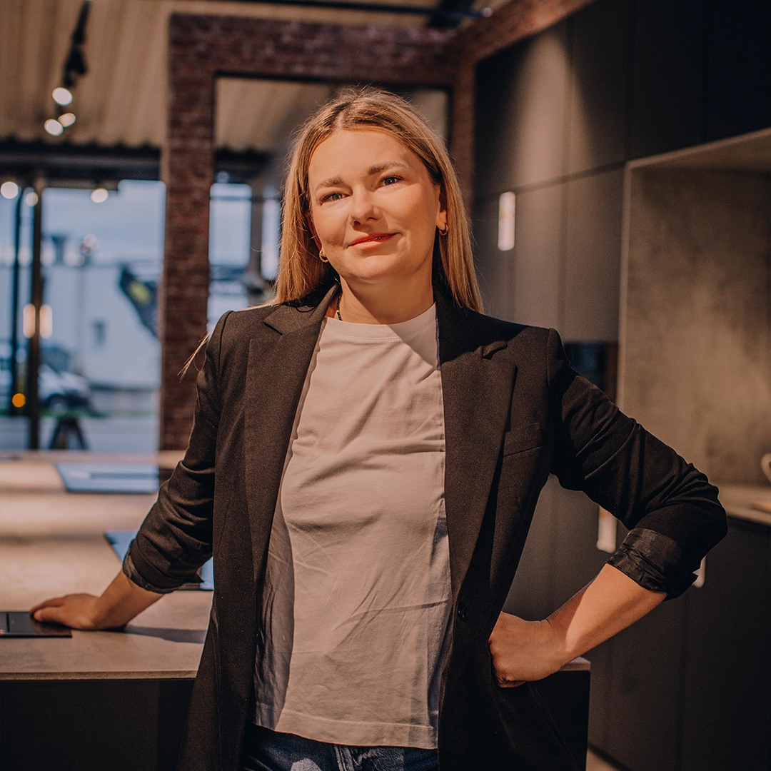 Eine Frau im schwarzen Blazer lächelt entspannt in einem modern gestalteten, gemütlichen Innenraum.