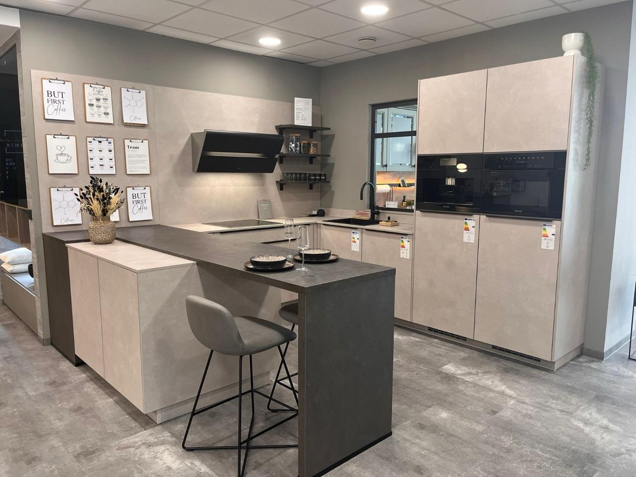 Moderne, aufgeräumte Ausstellungsküche in Grau und Beige mit Barhocker, goldener Vase und Wandbildern.