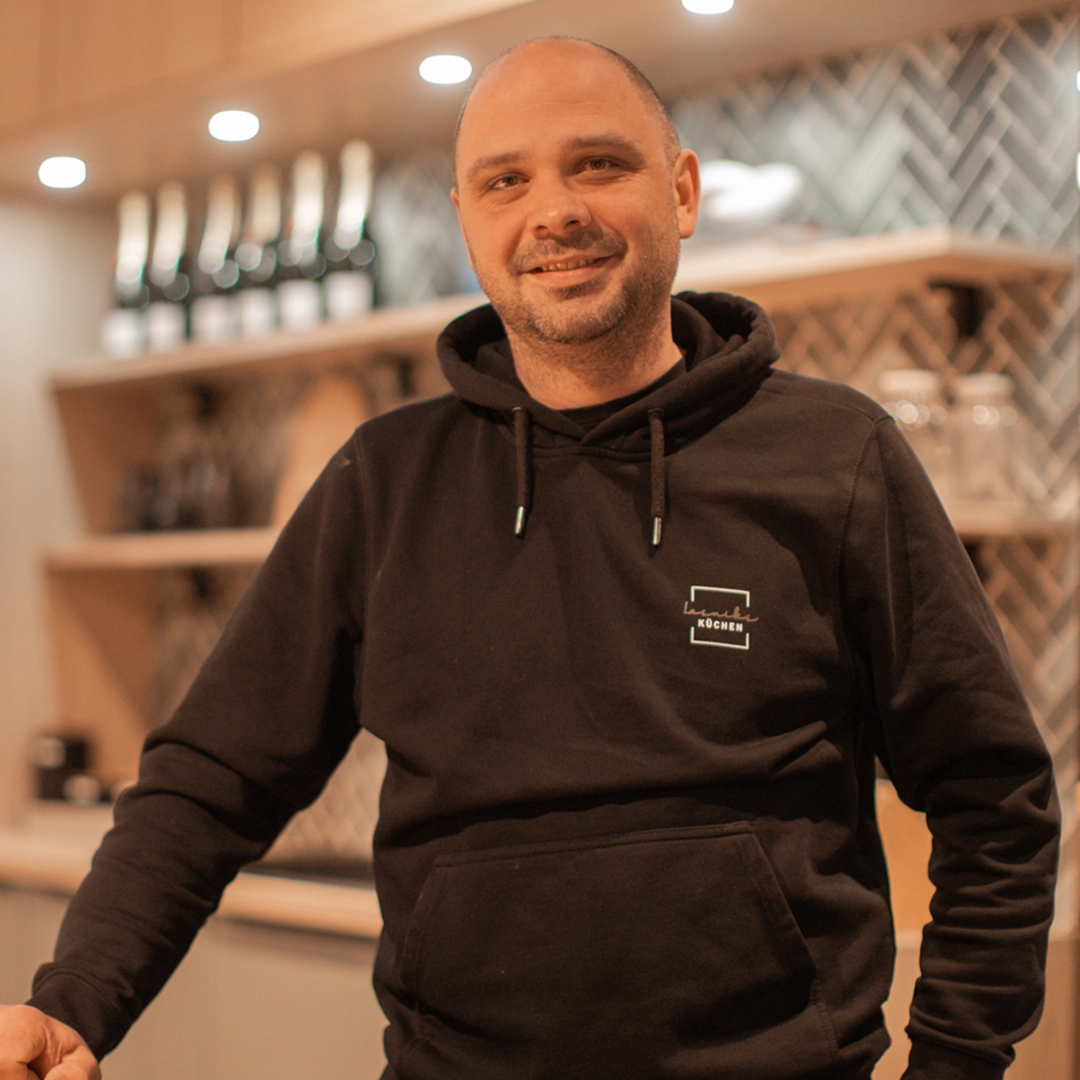 Eine Person im schwarzen Hoodie lächelt in einer warm beleuchteten, modernen Küche in die Kamera.