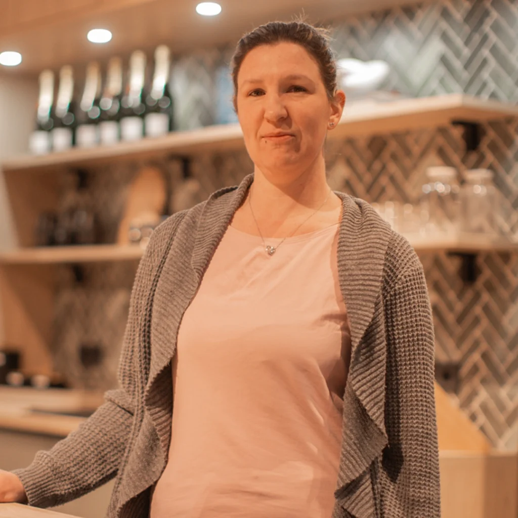 Frau in rosa T-Shirt und grauer Strickjacke steht entspannt in moderner Küche mit Regalen im Hintergrund.
