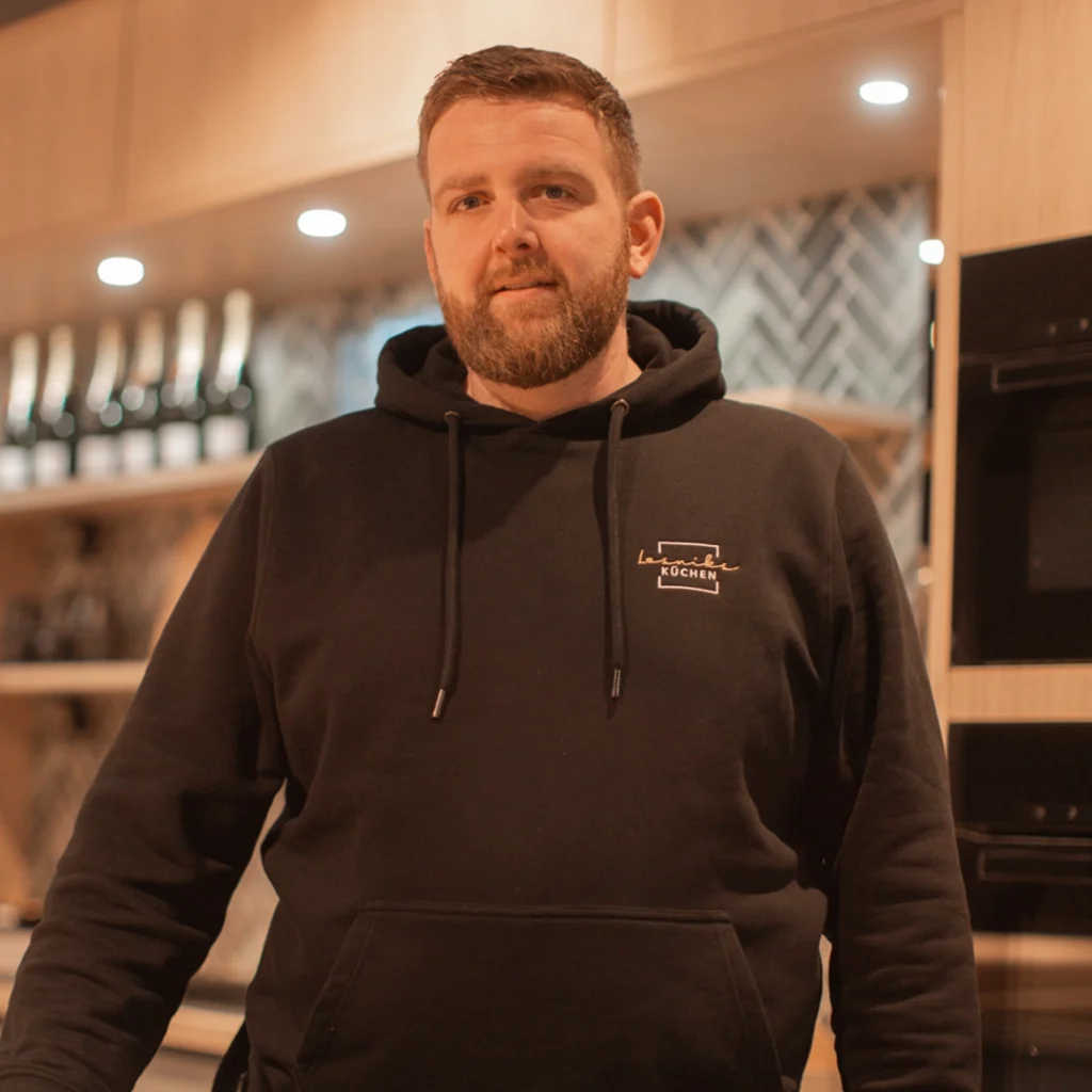 Person in schwarzem Hoodie steht in moderner Küche mit Regalen voller Flaschen, warm beleuchtet.