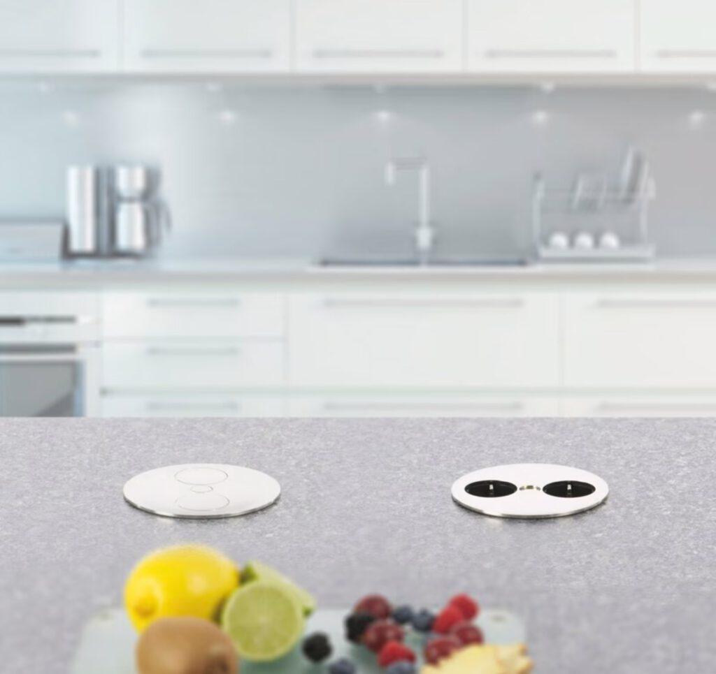 Moderne Küche mit grauer Arbeitsplatte, integriertem Steckdosen und buntem Obst auf einem Schneidebrett.