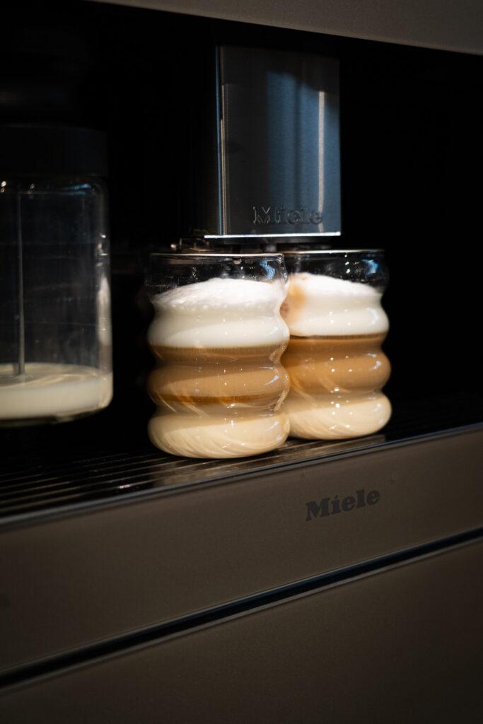 Miele Kaffeemaschine füllt zwei Gläser mit geschichtetem Latte Macchiato aus Milch, Kaffee und Schaum.