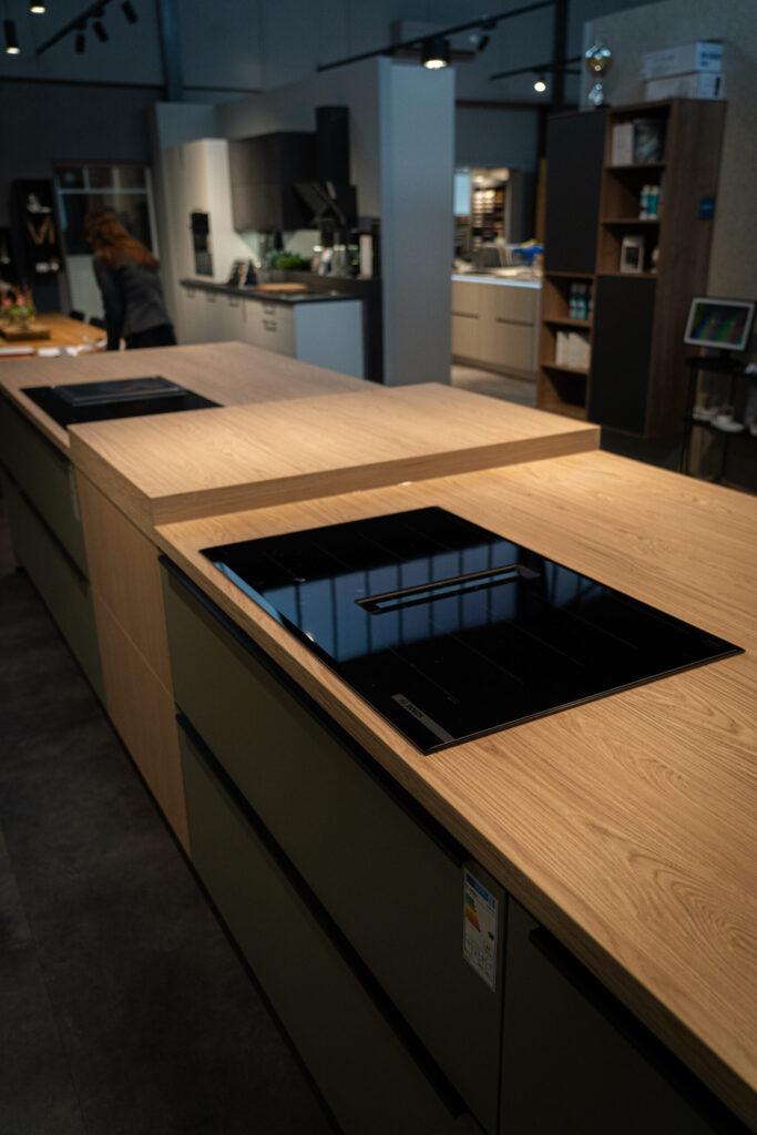 Moderne Küchenausstellung mit Holzinsel, Kochfeld und Person im Hintergrund, die Materialien betrachtet.