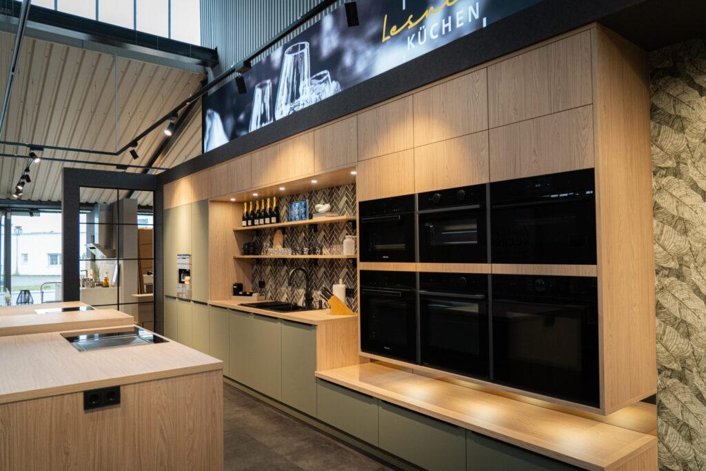 Moderne Ausstellungsküche mit grünen Schränken, Holzdetails und dekorativen Regalen in warmem Licht.