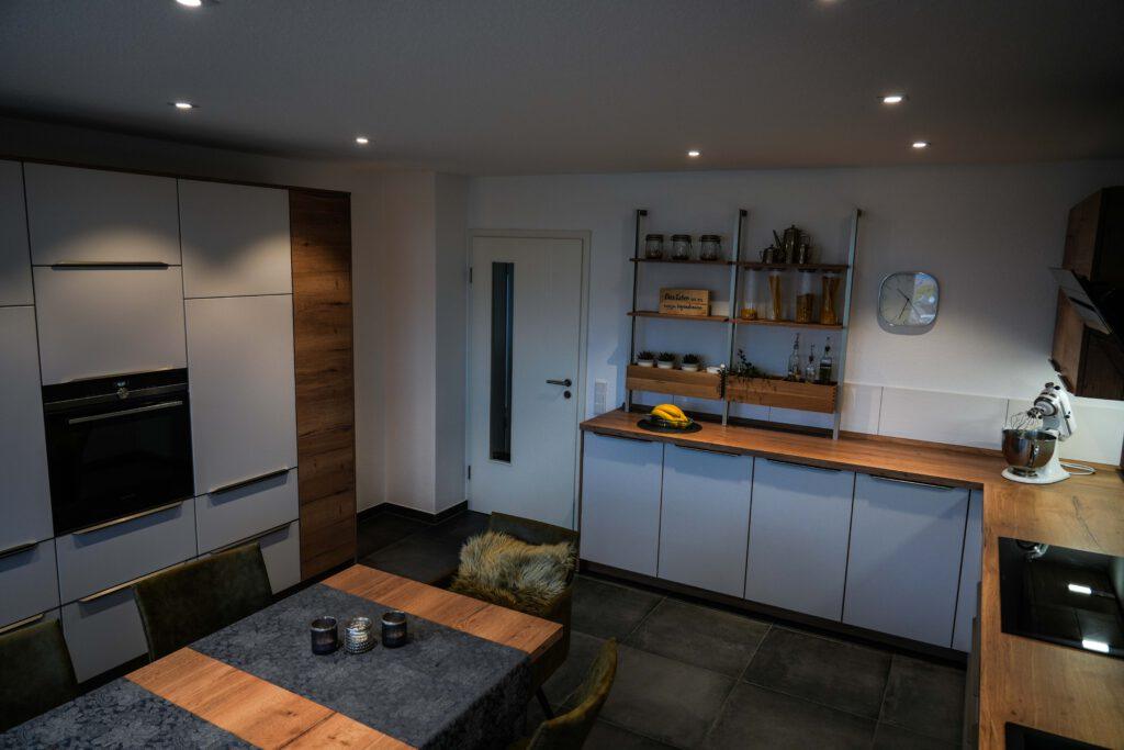 Moderne Küche mit weißen und holzfarbenen Schränken, Esstisch, Regalen und gelben Paprikas.