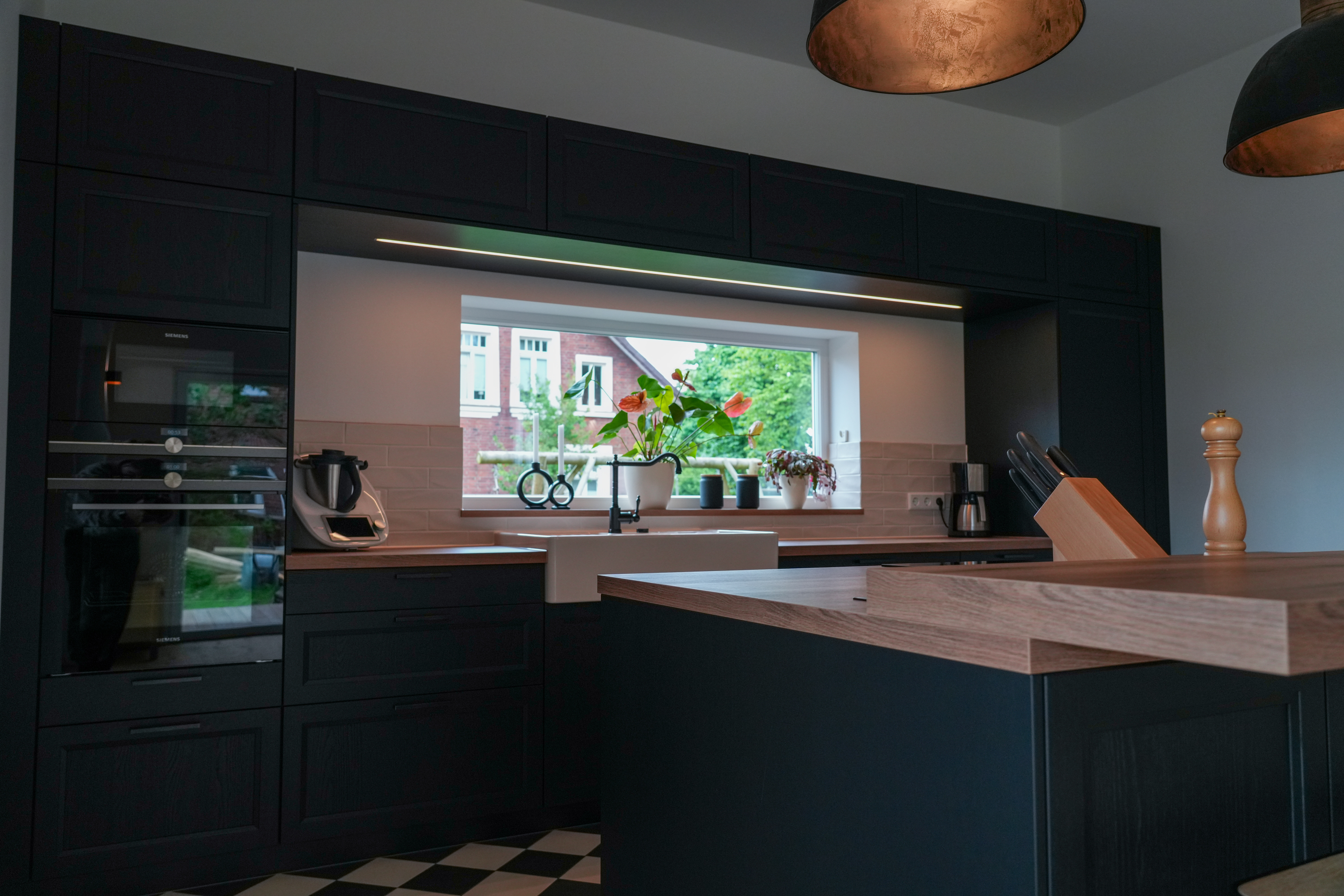 Moderne Küche mit dunklen Schränken, Holzarbeitsplatte, großem Fenster und Pflanzen auf der Fensterbank.