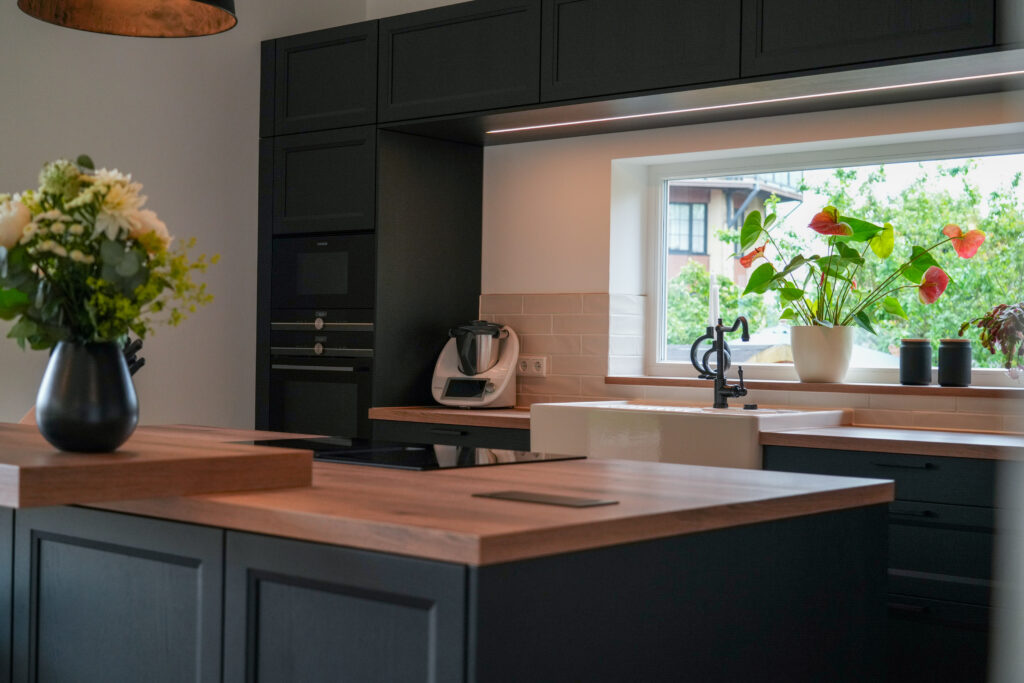 Moderne Küche mit dunklen Schränken, Holzarbeitsplatte, Pflanzen am Fenster und einem Blumenstrauß.