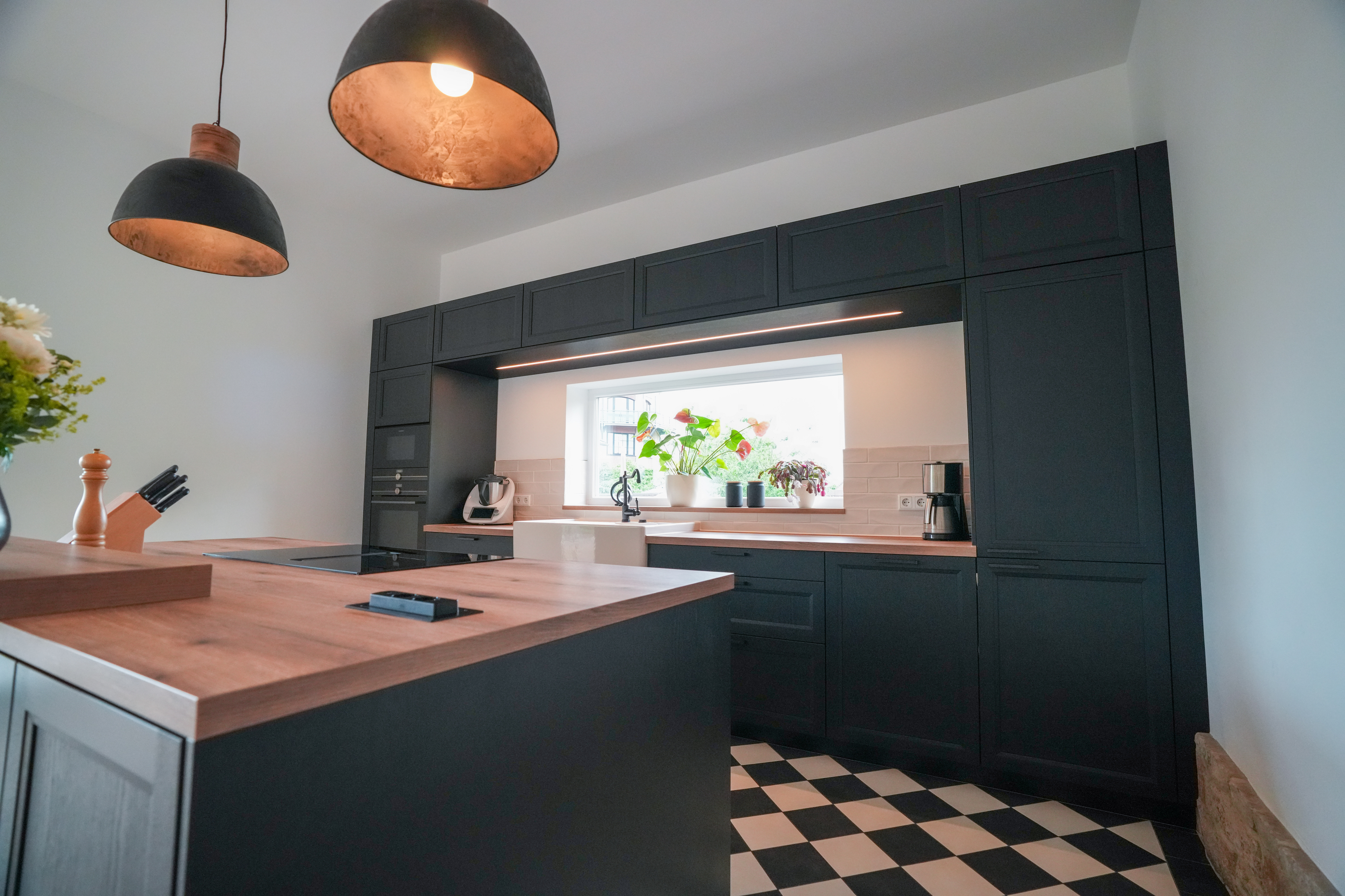 Moderne Küche mit dunklen Schränken, Holzarbeitsplatte, Hängelampen und großen Fenstern.