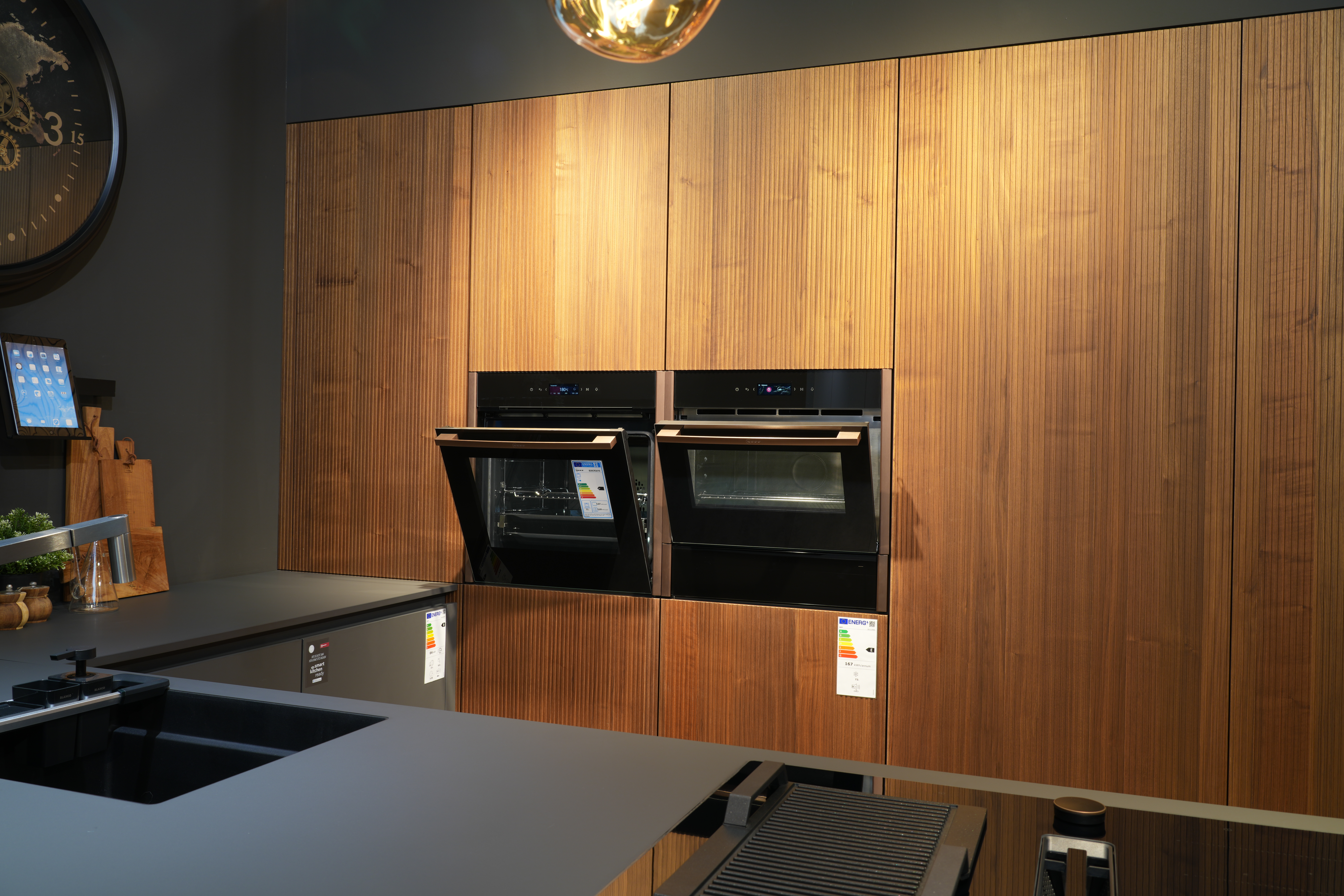 Moderne Küche mit Holzschränken, zwei schwarzen Backöfen, dunkler Arbeitsplatte und runder Wanduhr.