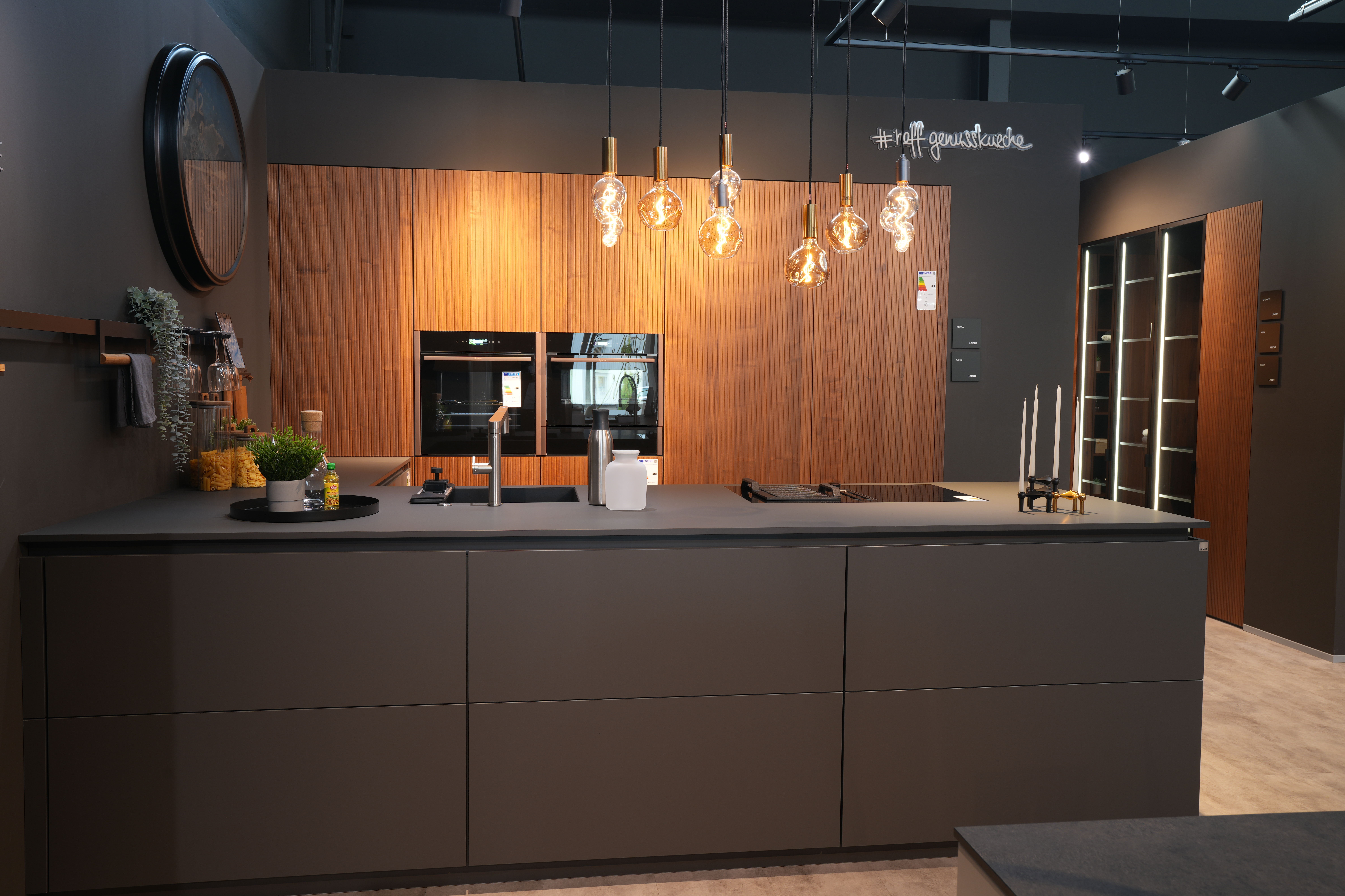 Moderne Küche mit dunkler Insel, hängenden Glühbirnen und Holzwand, warm beleuchtet und stilvoll.