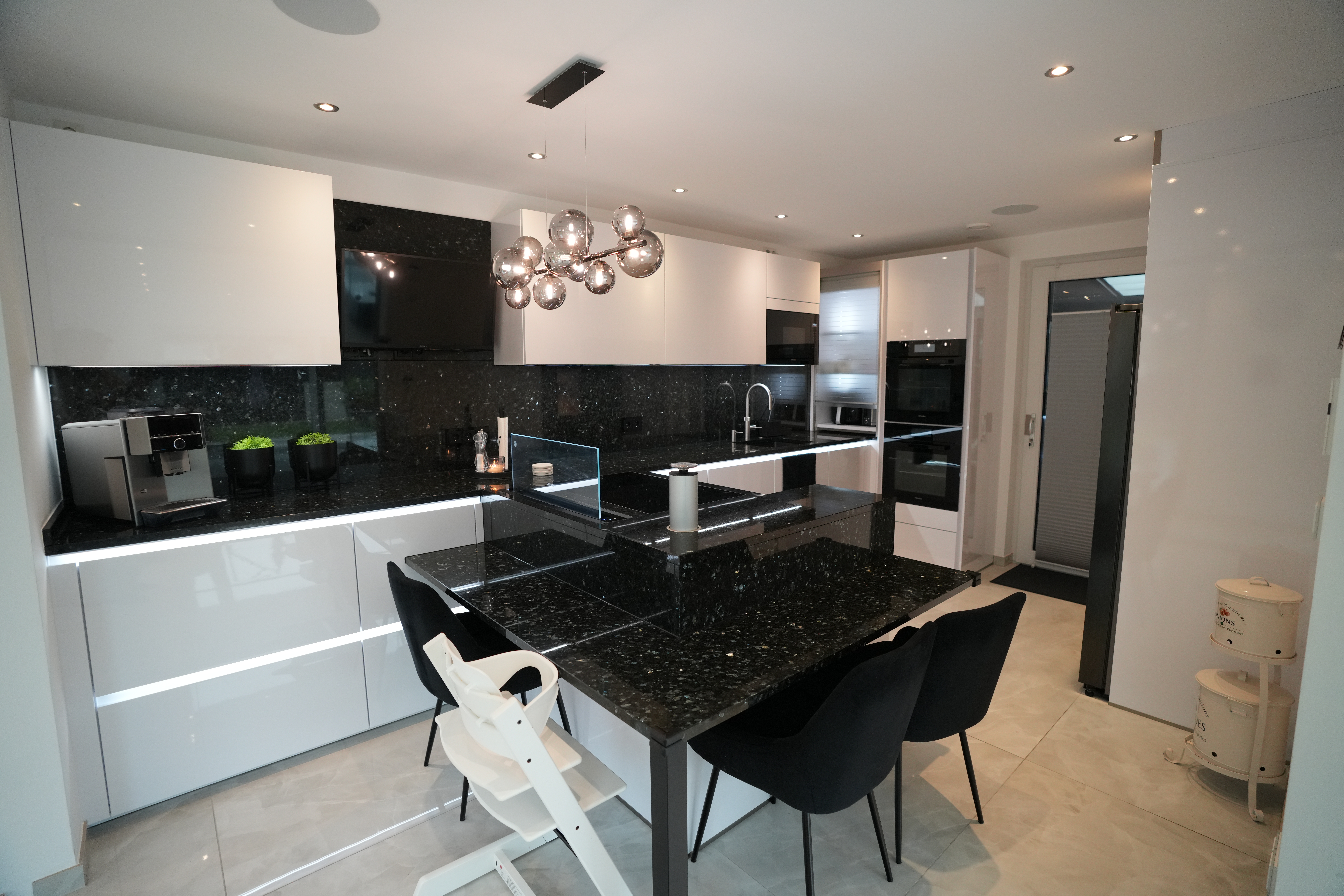 Moderne Küche mit weißen Schränken, dunklen Arbeitsflächen und stilvoller Beleuchtung. Keine Personen sichtbar.