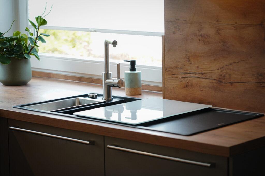 Moderne Küchenecke mit Edelstahlspülbecken, Wasserhahn, Seifenspender und grüner Pflanze.