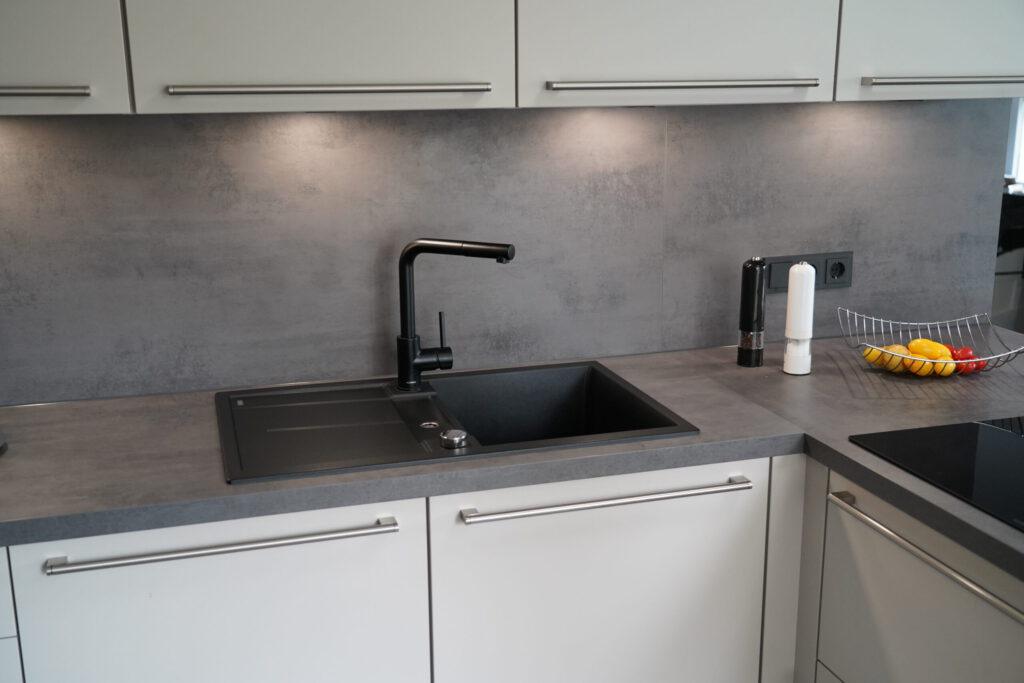 Moderne Küche mit schwarzer Spüle, grauer Arbeitsplatte, weißen Schränken und buntem Obstkorb.