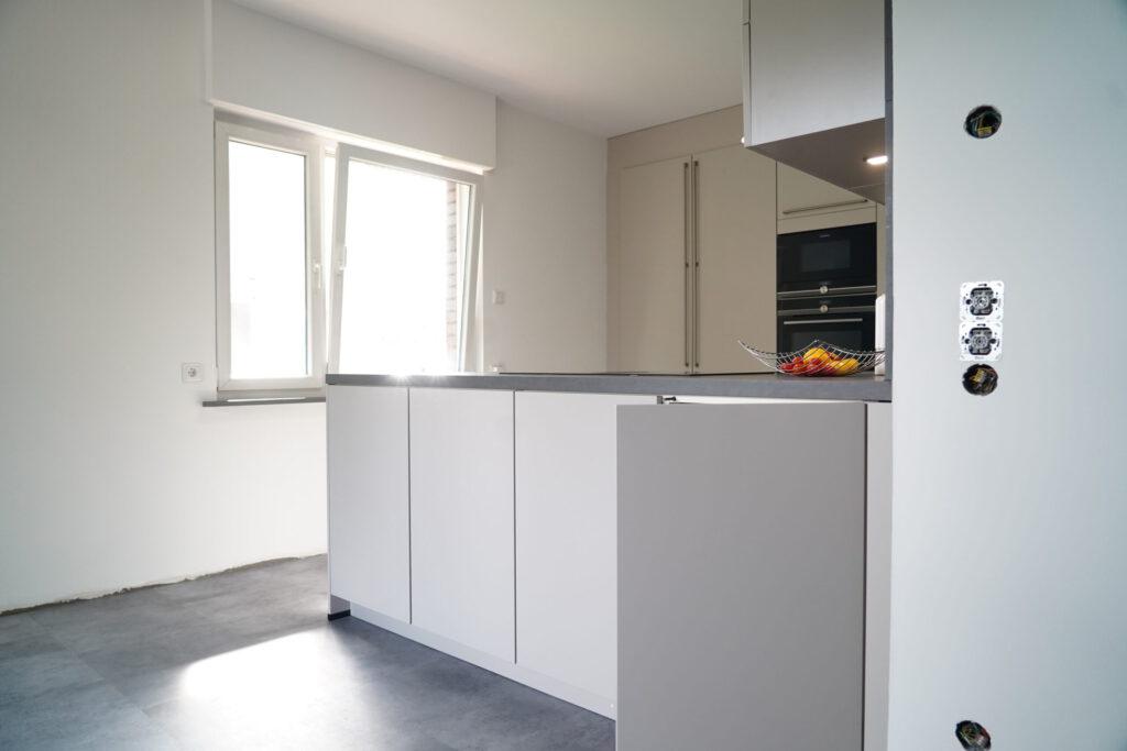 Moderne, minimalistische Küche mit grauer Insel, Obstschale und natürlichem Licht durch offenes Fenster.