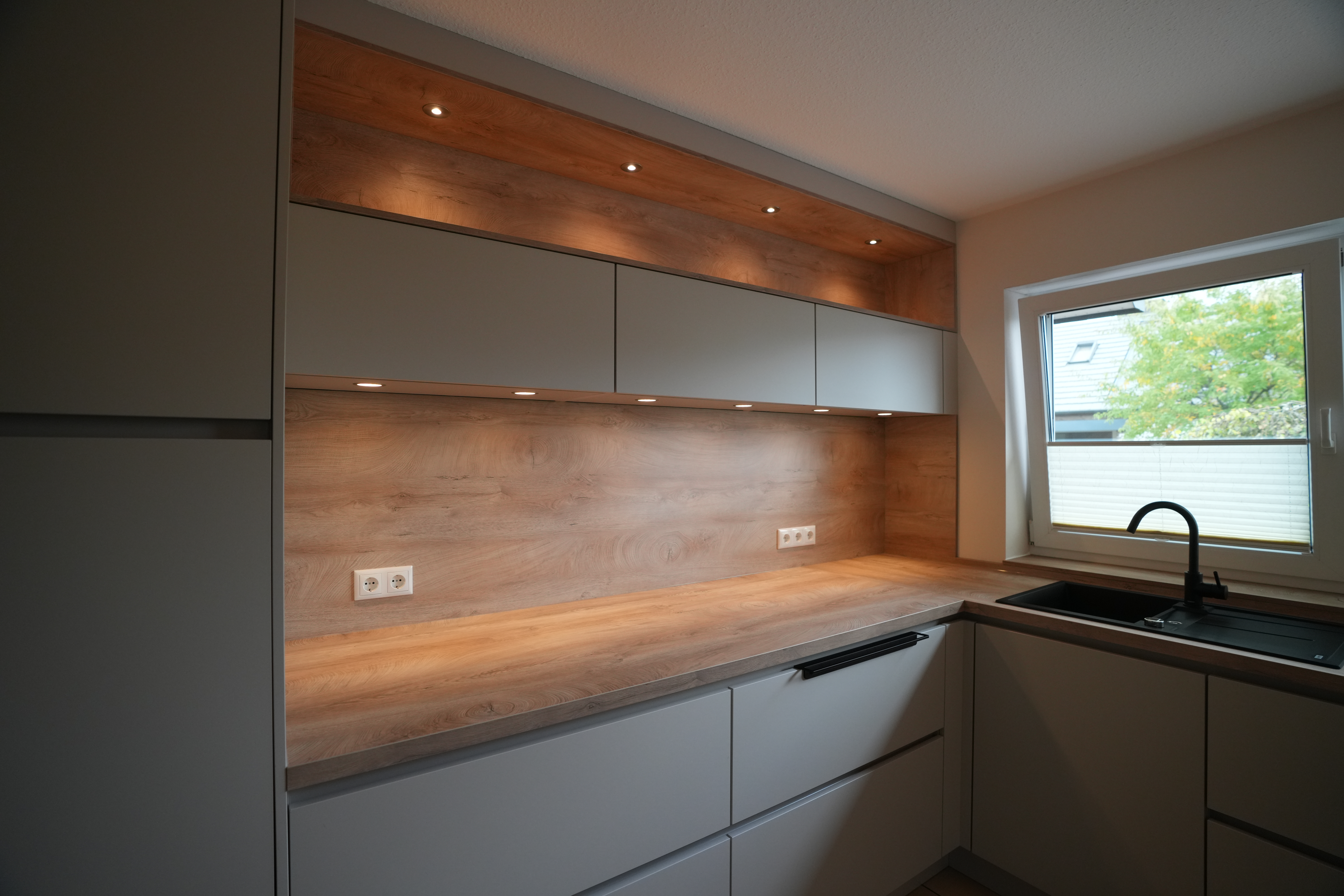 Moderne Küche mit grauen Schränken, Holzarbeitsplatte, schwarzer Spüle und Fenster mit Außenblick.