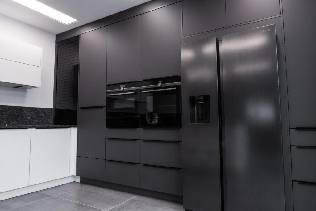 Moderne Küche mit grauen Schränken, schwarzer Arbeitsplatte, großem Kühlschrank und zwei Einbauöfen.