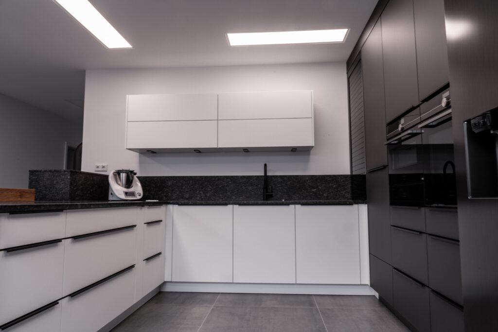 Moderne, unbenutzte Küche mit weißen und grauen Schränken, dunkler Arbeitsplatte und eingebauten Geräten.