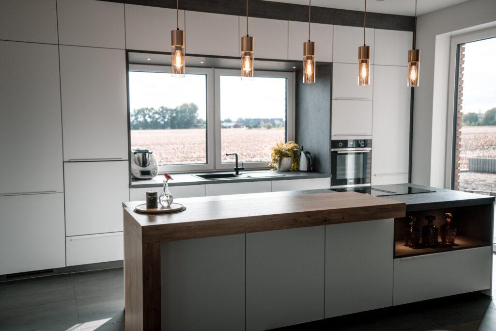 Moderne, aufgeräumte Küche mit Holzinsel, warmem Licht und Blick auf ein Feld durch ein großes Fenster.