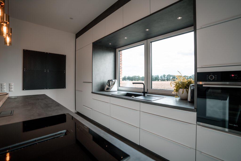 Moderne, minimalistische Küche mit weißen Schränken, viel Licht und einer Pflanze auf der Fensterbank.