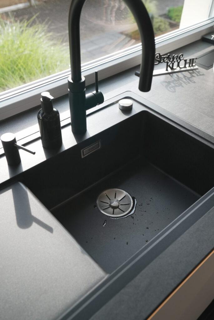 Moderne schwarze Küchenspüle mit Wasserhahn, Seifenspender und Fenster im Hintergrund.