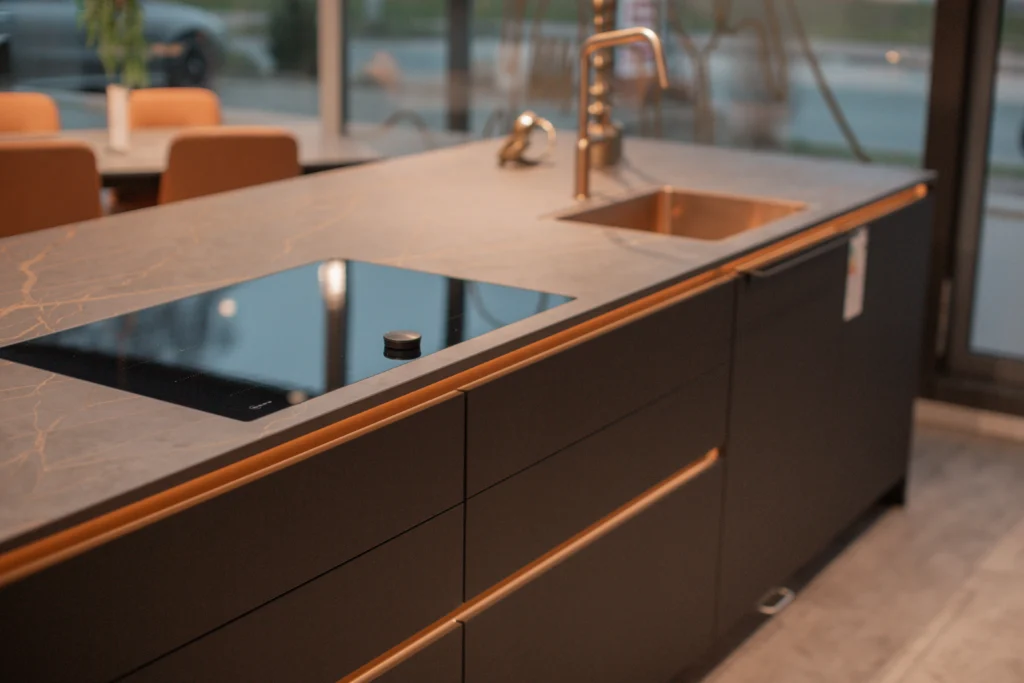 Moderne Küche mit grauer Arbeitsplatte, schwarzem Kochfeld, metallischem Spülbecken und orangen Stühlen.