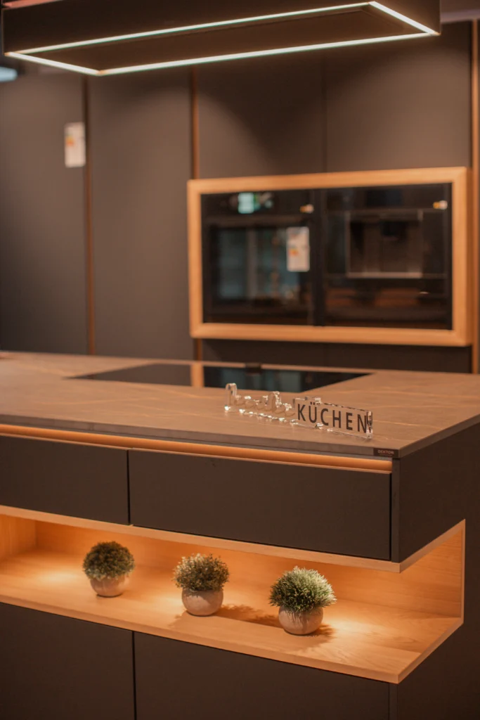 Moderne Küche mit dunkler Arbeitsplatte, Holzrahmen, Pflanzen und eingebauten Öfen im Hintergrund.