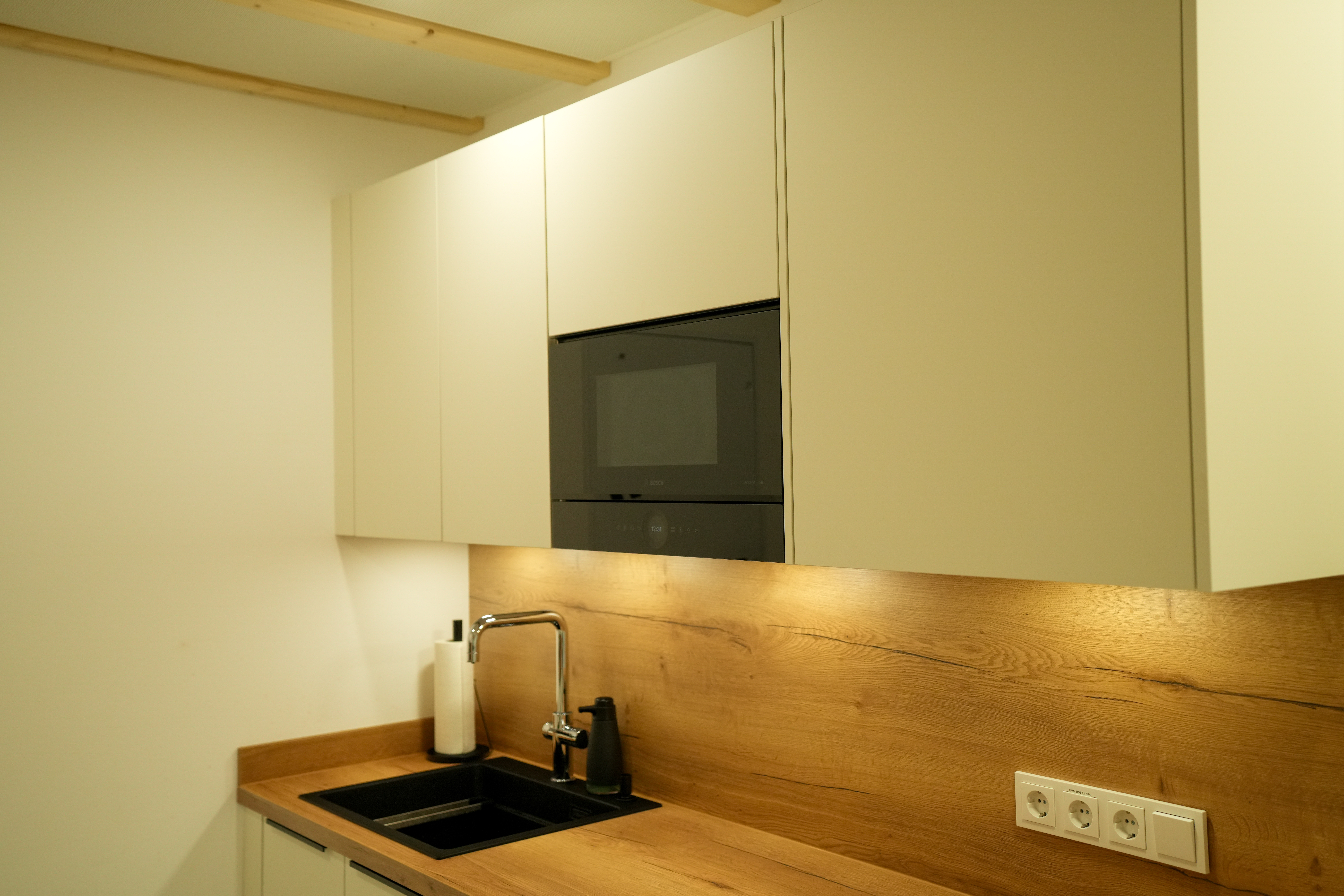 Moderne Küche mit beigen Schränken, schwarzer Spüle und Holzarbeitsplatte in warmem Licht.
