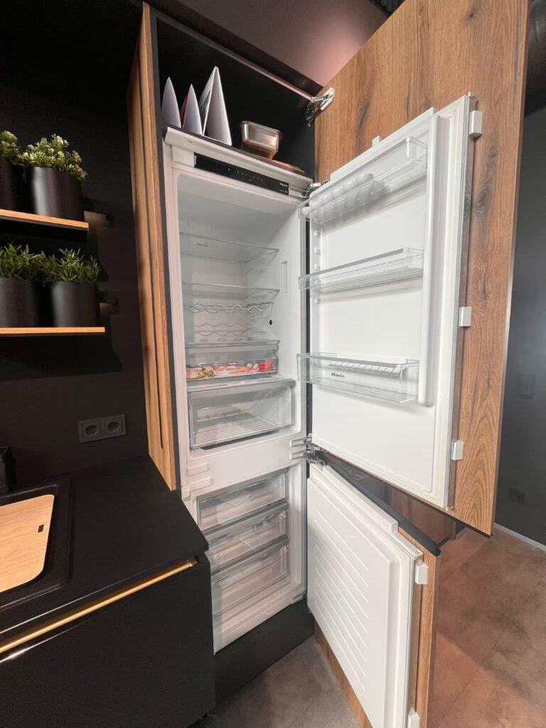 Geöffneter Einbaukühlschrank in moderner Küche mit Holzschränken und schwarzen Regalen mit Pflanzen.