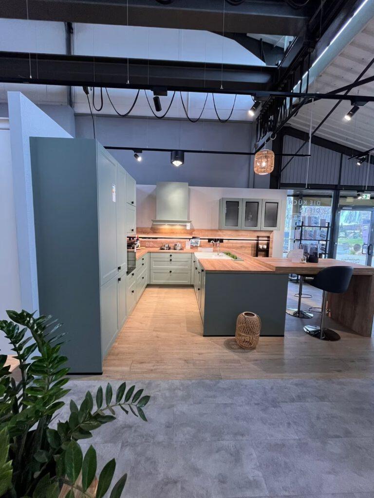 Moderne, unbenutzte Küche in sanftem Grün mit Holzarbeitsplatte, Barhocker und großer Pflanze.