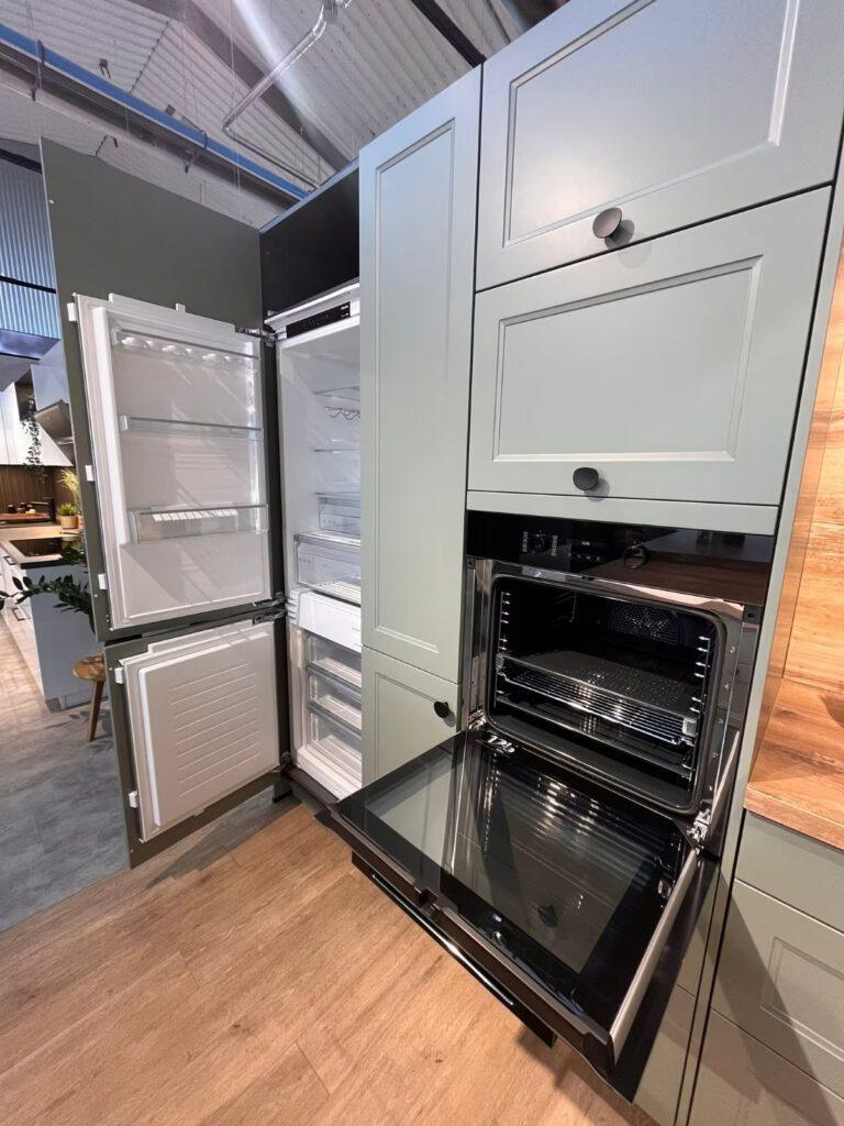 Moderne Küche mit offenem Kühlschrank und Backofen, graugrünen Schränken und Holzoptik-Boden.
