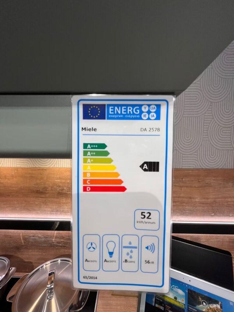 Energieeffizienzlabel für Miele DA 2578, Klasse A, 52 kWh/Jahr, vor Küchengeräten im Hintergrund.