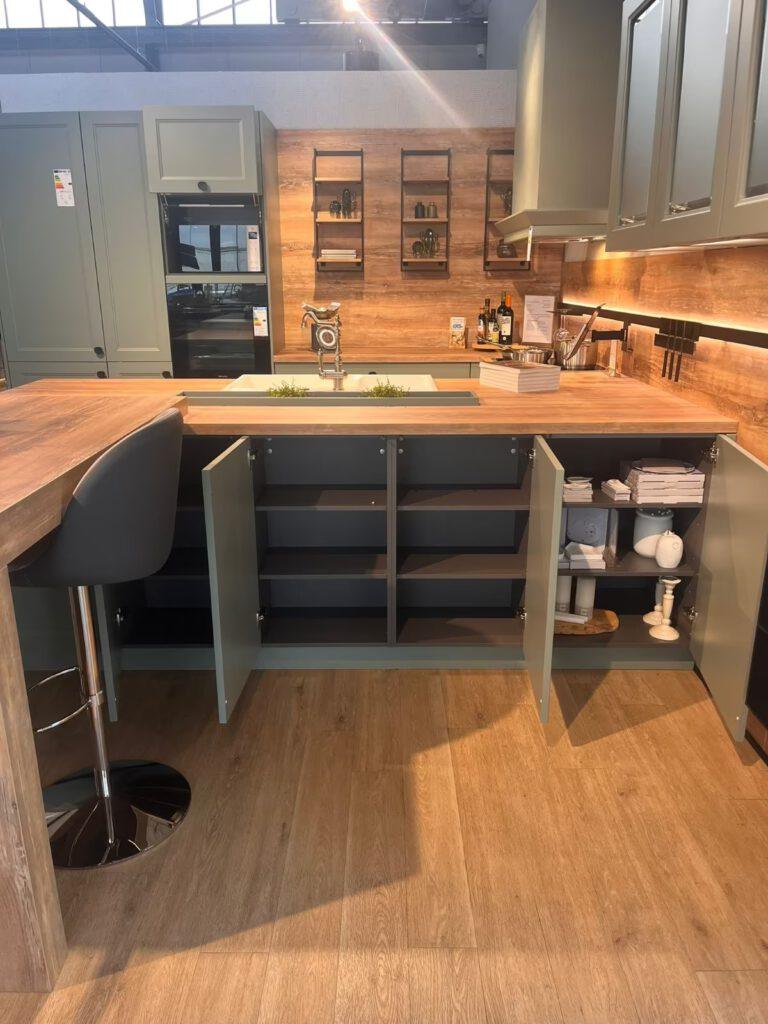 Moderne Ausstellungsküche in sanftem Graugrün mit Holzarbeitsplatte, Barhocker und dekorativen Regalen.