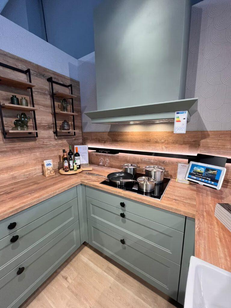Moderne Küche mit grünen Schränken, Holzarbeitsplatte, Kochfeld, Dunstabzugshaube und offenen Regalen.