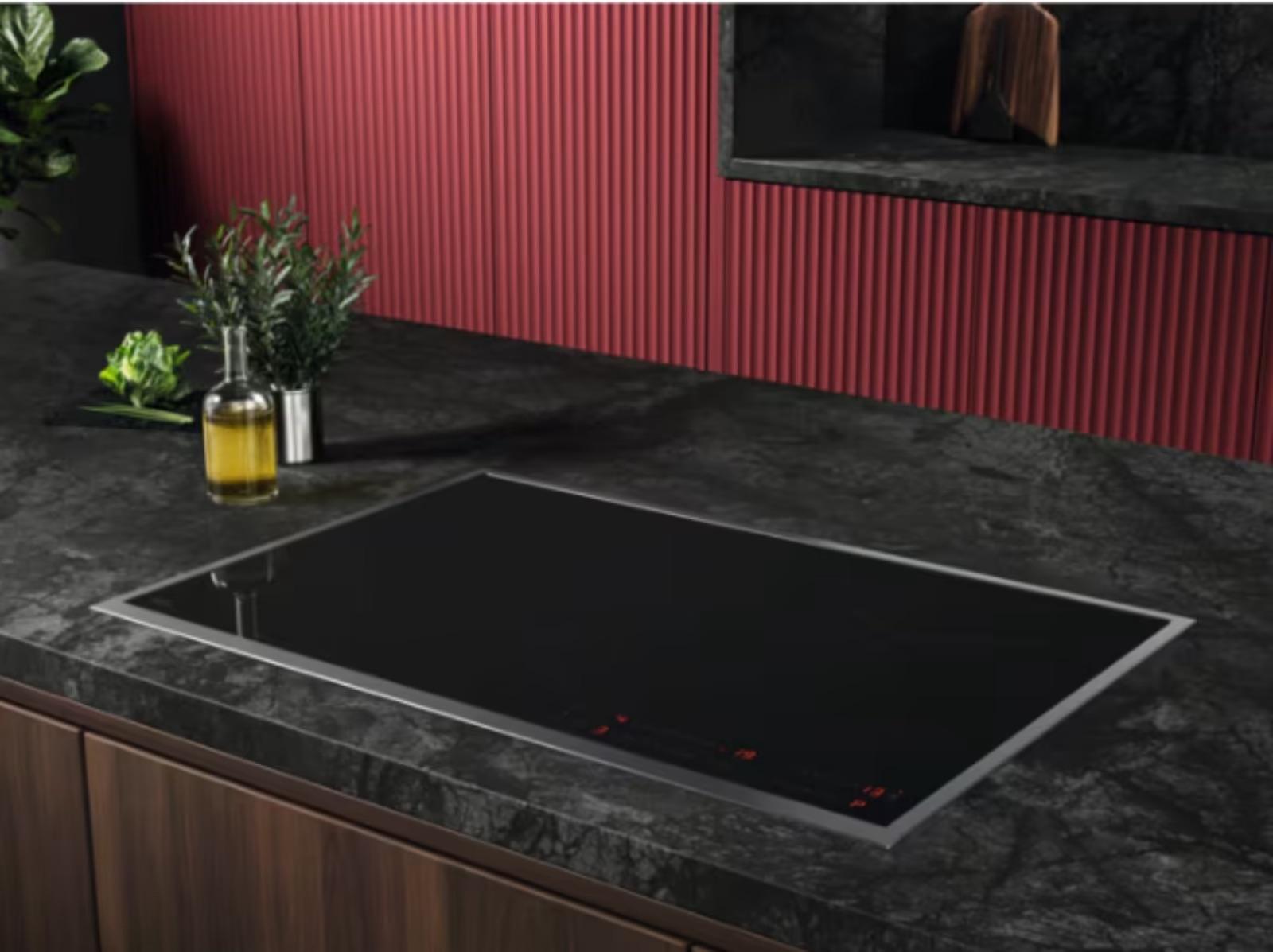 Moderne Küche mit schwarzem Induktionskochfeld, Ölflasche, Kräutern und grünem Salat auf dunkler Arbeitsplatte.