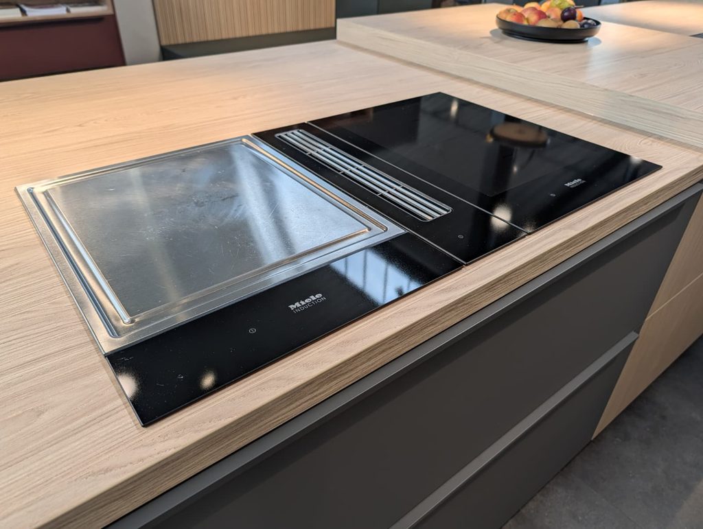 Moderne Küche mit hellem Holz, schwarzem Induktionskochfeld und einer Schale bunter Früchte.