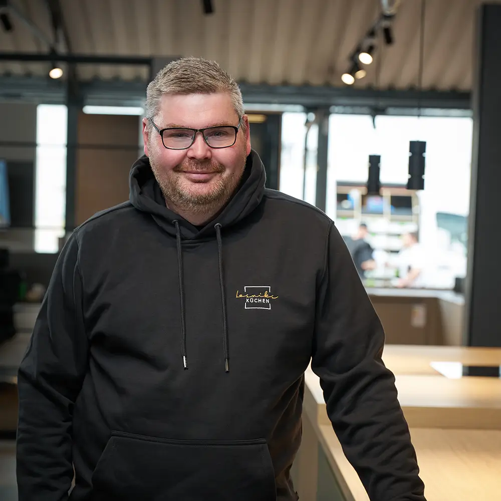 Person im dunklen Hoodie steht entspannt in modernem Raum, schaut freundlich in die Kamera.