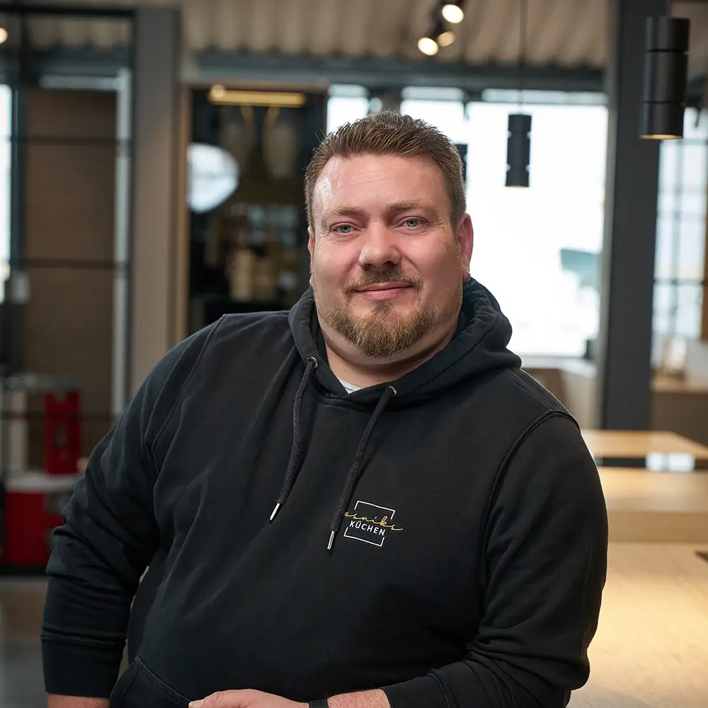 Eine Person lehnt sich entspannt in einem modernen Raum an, lächelt in die Kamera; warme, neutrale Farben.