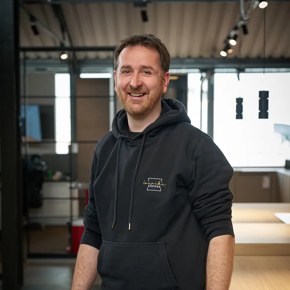 Person im dunklen Hoodie lächelt in modernem Büro mit industrieller Decke und Holztisch.