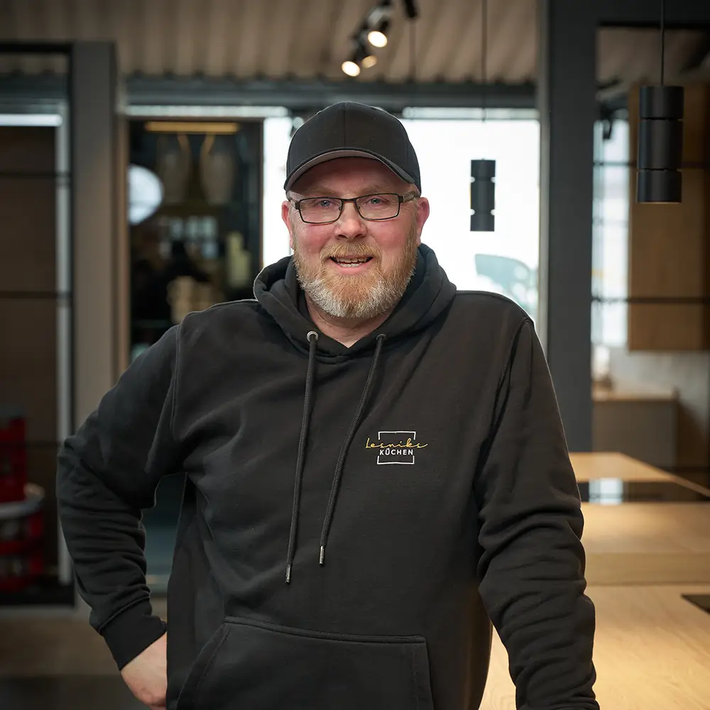 Ein Mann in schwarzem Hoodie und Kappe lächelt in einem modernen, stilvollen Innenraum in die Kamera.
