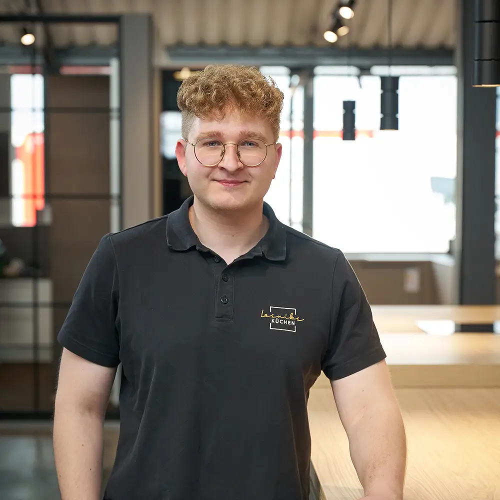 Ein junger Mann im schwarzen Polohemd lächelt freundlich in einem modernen, minimalistischen Raum.