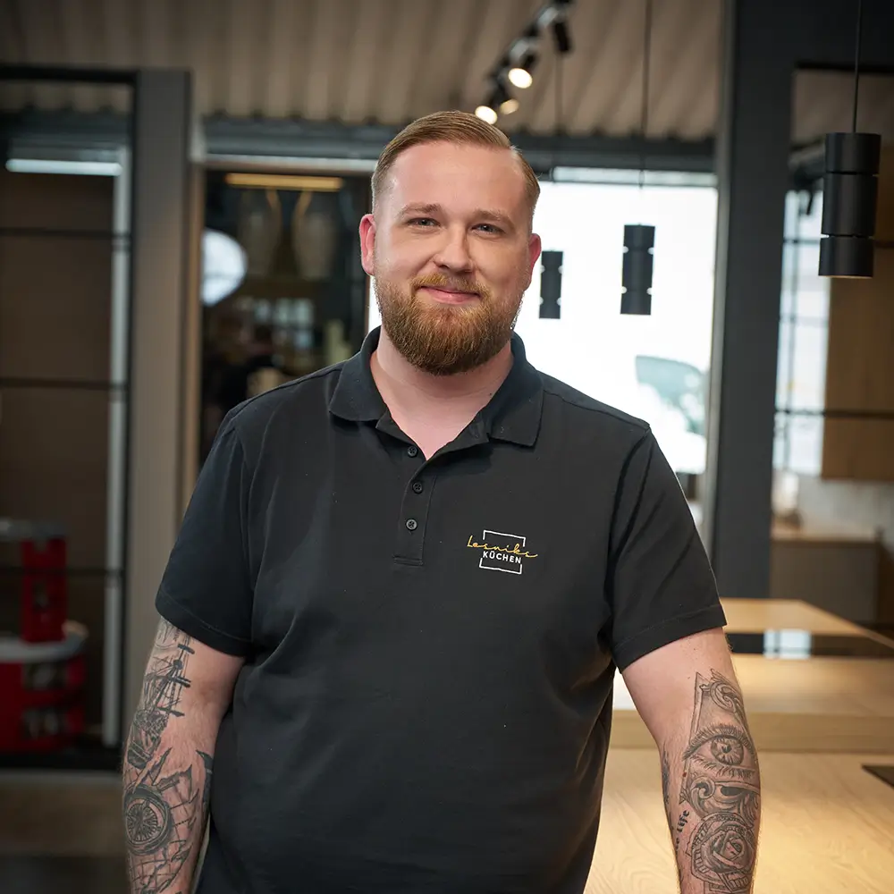 Ein Mann im schwarzen Polohemd mit Logo lächelt in einem modernen, stilvollen Innenraum in die Kamera.
