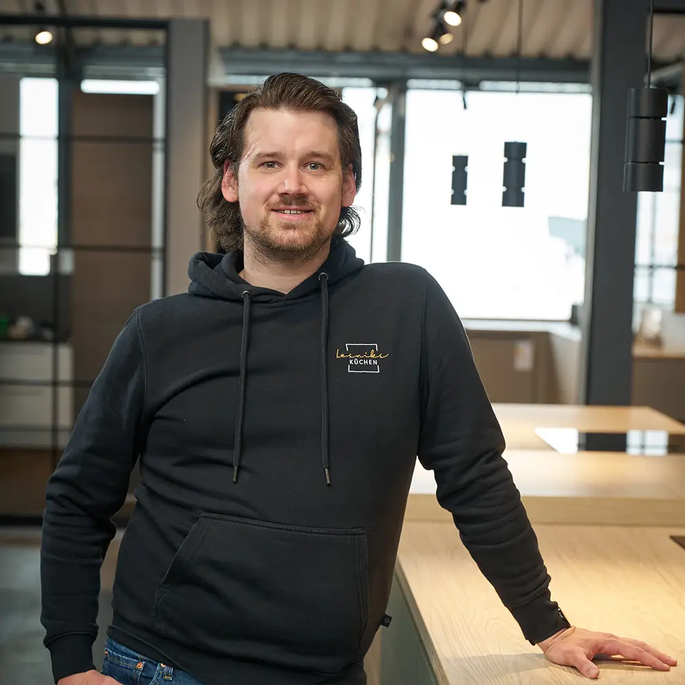Ein Mann in schwarzem Hoodie lehnt lächelnd an einem Holztisch in moderner, gut beleuchteter Umgebung.