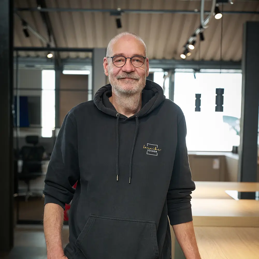 Eine Person im dunklen Hoodie lächelt in die Kamera, vor einem modernen, neutralen Hintergrund.