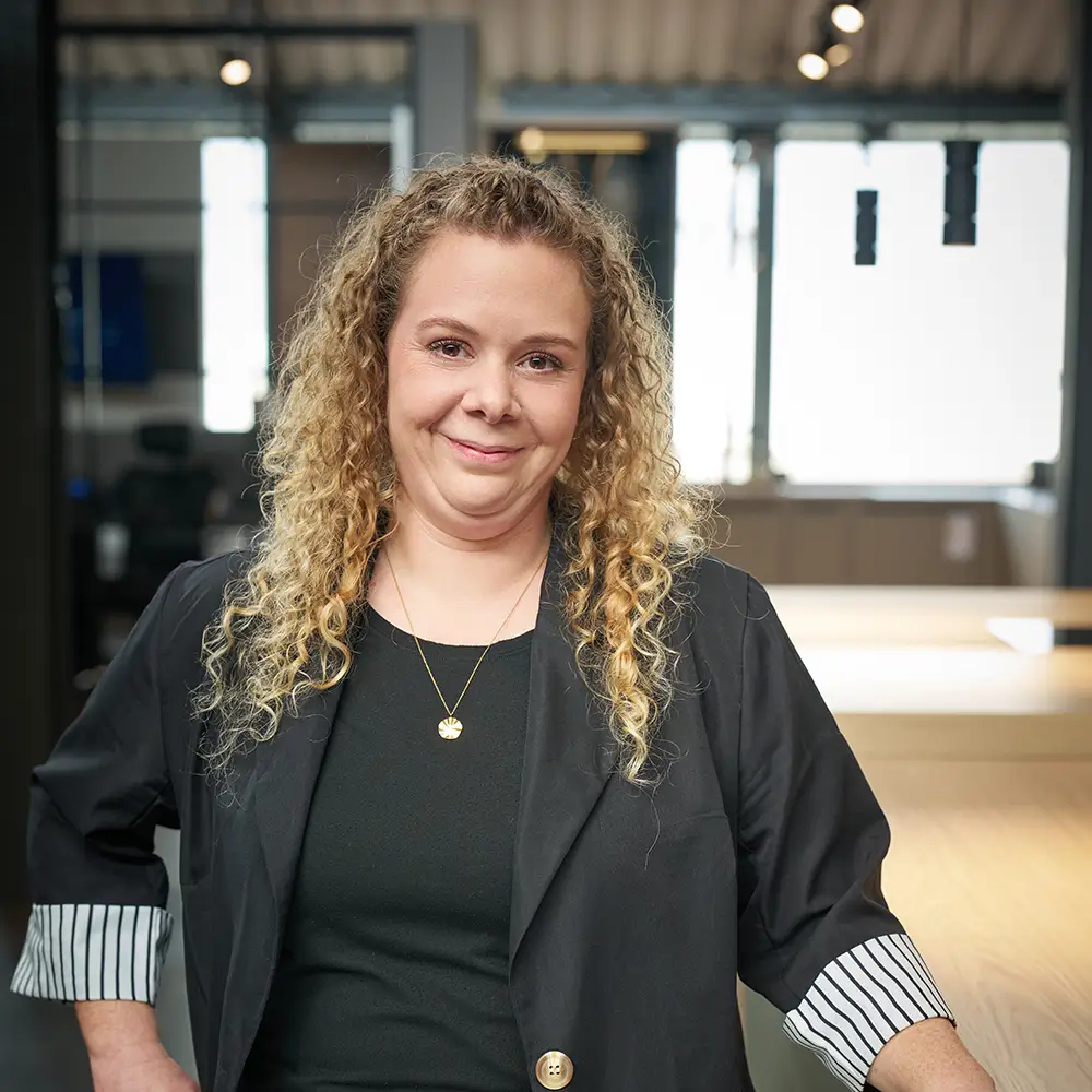 Eine lächelnde Person in einem schwarzen Blazer steht in einem hellen, modernen Büro.