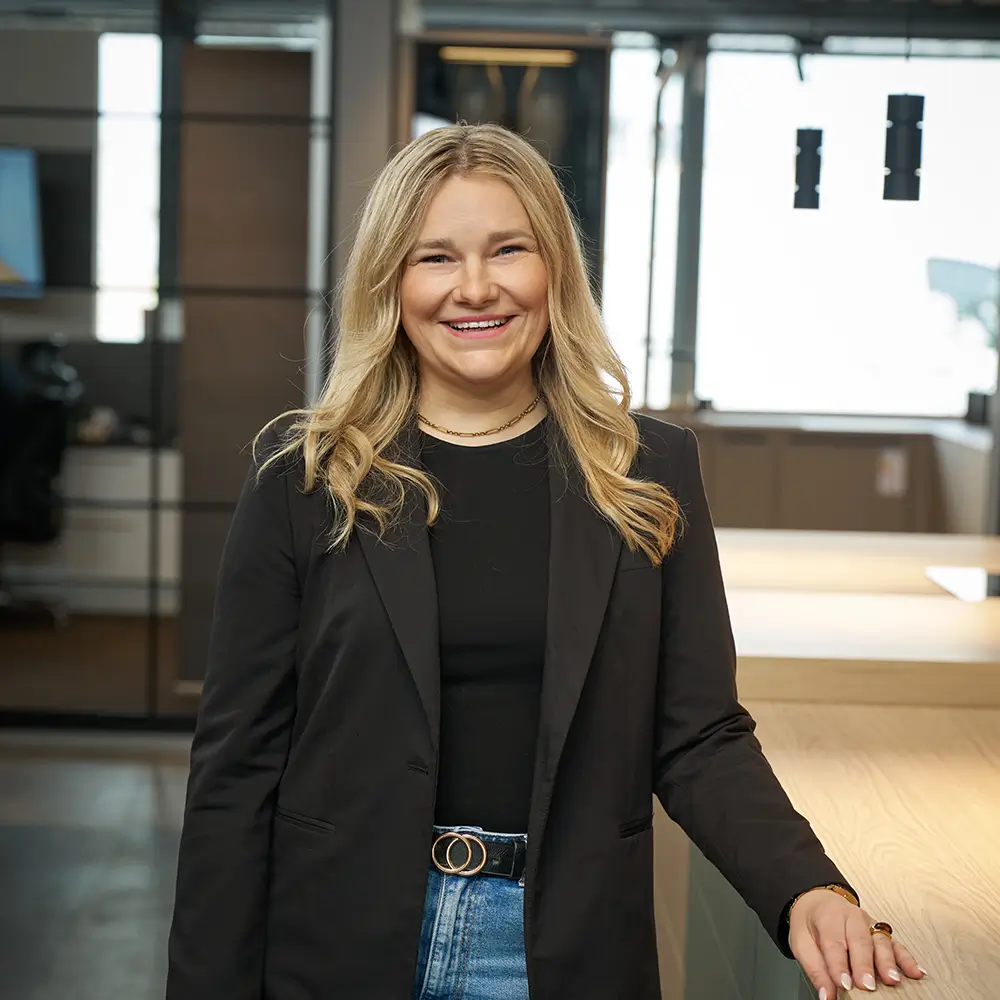Eine lächelnde Frau im schwarzen Blazer steht in einem modernen Büro mit Glaswand und Holztisch.