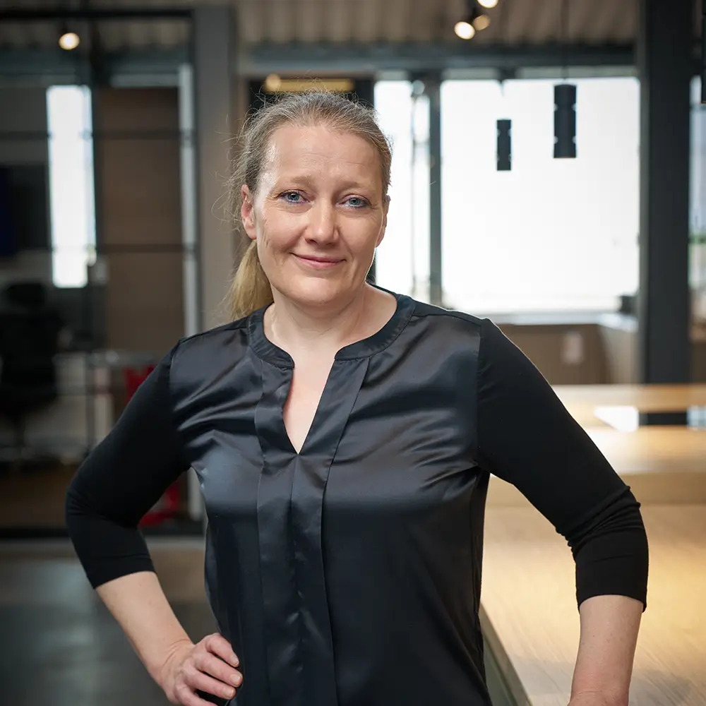Frau in schwarzem Oberteil steht selbstbewusst in modernem Büro, schaut freundlich in die Kamera.